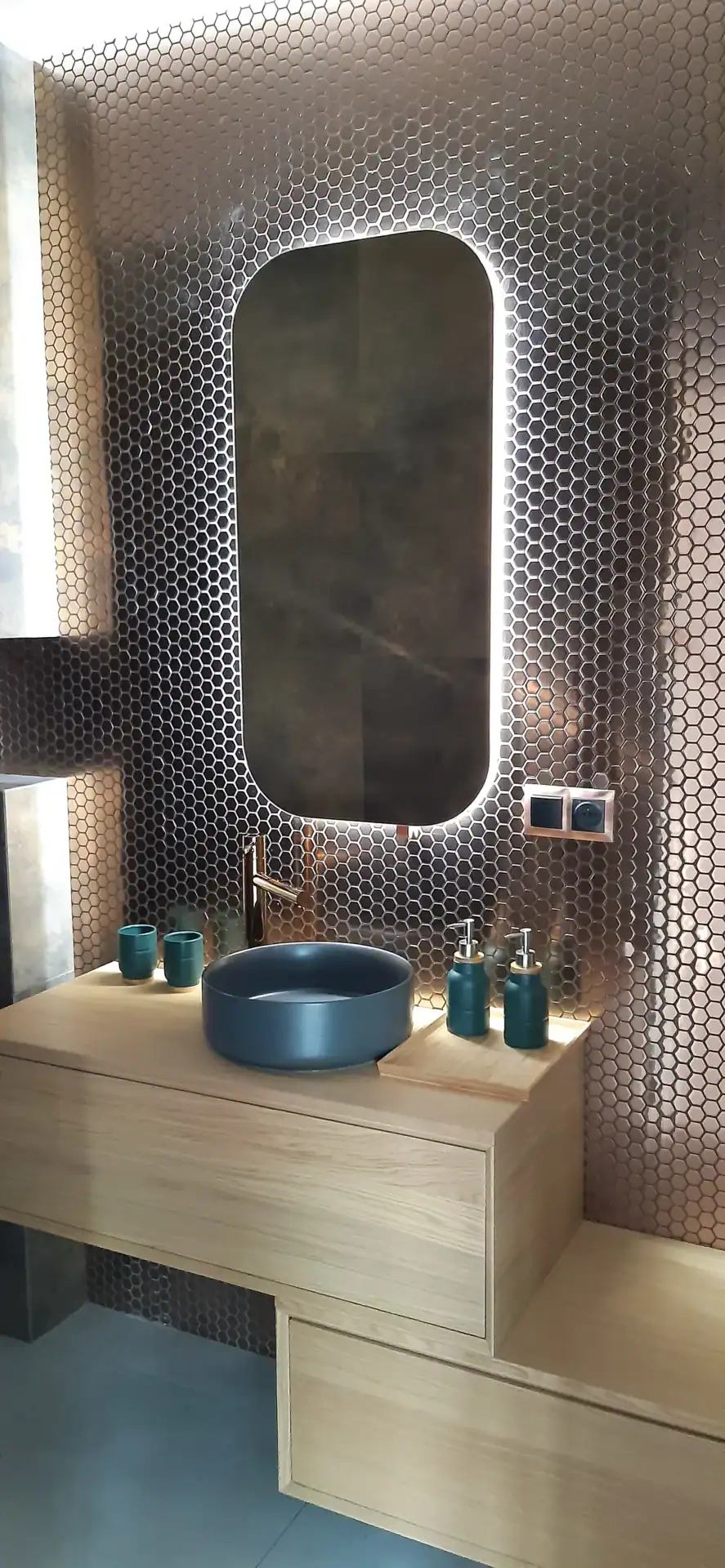 Nowoczesna łazienka z drewnianą toaletką 20210711_172439-scaled-1.webp, owalną niebieską okrągłą umywalką, pasującymi dozownikami mydła, dużym owalnym podświetlanym lustrem i ścianą ozdobioną małymi okrągłymi metalicznymi płytkami.