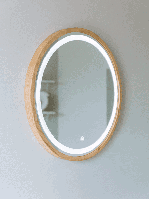 Pitagoras-Perfection-Slim-LED1.png to okrągłe, montowane na ścianie lustro z drewnianą ramą i wbudowanym oświetleniem LED, pokazane jako odbicie minimalistycznego pokoju z białymi ścianami i subtelnymi półkami w tle.