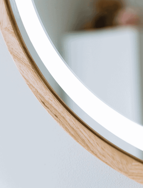 Zbliżenie na okrągłe lustro Pitagoras-Perfection-Slim-LED2.png z drewnianą ramą i podświetlanym białym obramowaniem LED, odbijające rozmyte kształty w minimalistycznym, jasnym pomieszczeniu.