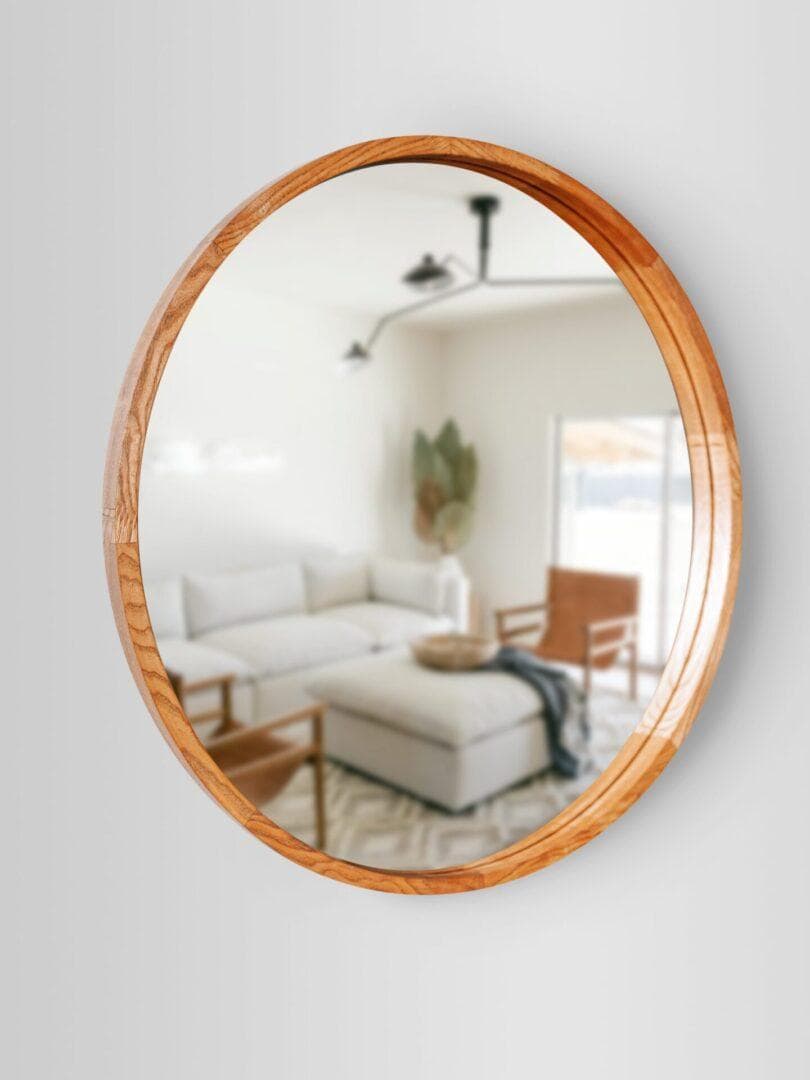 Lustro koniak2-scaled-1.jpg w drewnianej ramie odbija nowoczesny salon z jasną sofą, pufem, drewnianymi krzesłami, roślinami i lampą sufitową - wszystko delikatnie rozmazane w odbiciu.