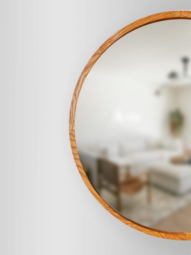 Koniak3-scaled-1.jpg, dekoracyjne lustro w drewnianej ramie, wisi na białej ścianie i odbija miękką, rozmytą scenę nowoczesnego salonu z neutralnymi meblami i akcentami dekoracyjnymi.