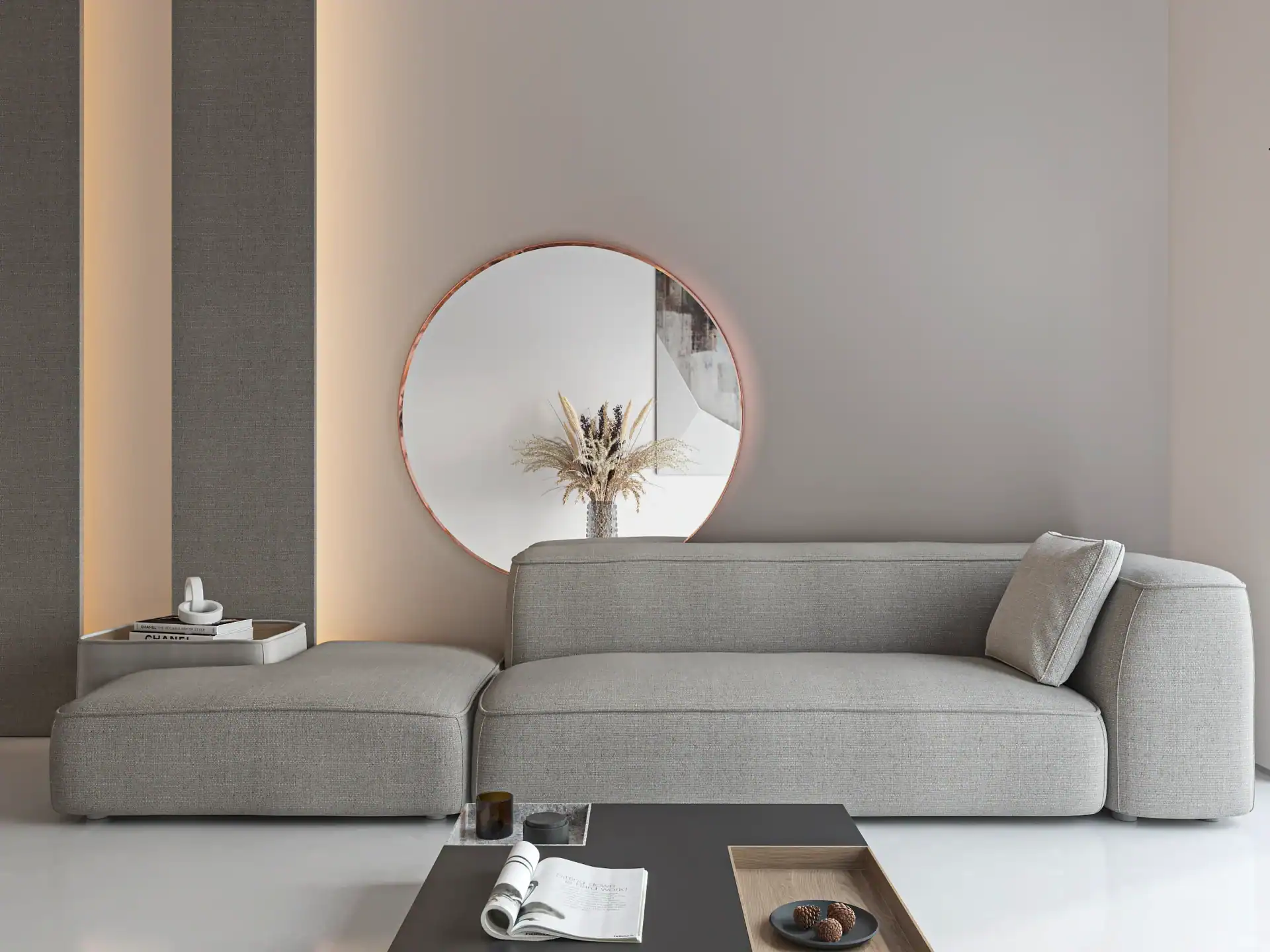 W nowoczesnym salonie znajduje się jasnoszara sofa segmentowa miedz-scaled-1.webp, okrągłe lustro ścienne i neutralny wystrój inspirowany stylem Rama. Czarny stolik kawowy z czasopismami i przekąskami uzupełniają suszone kwiaty w wazonie obok lustra.