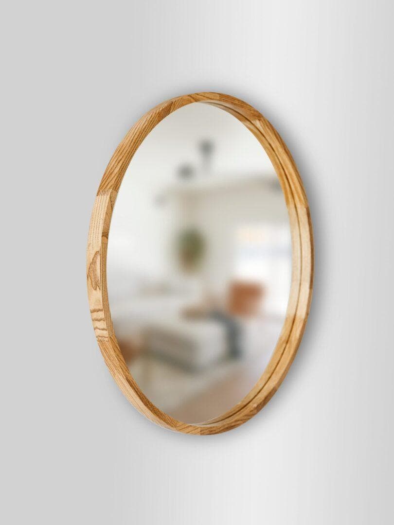 Lustro popiel2-scaled-1.jpg, oprawione w drewno, wisi na jasnoszarej ścianie i odbija rozmazany widok przytulnego, jasnego pokoju z sofą, dużym oknem i gustownym wystrojem.