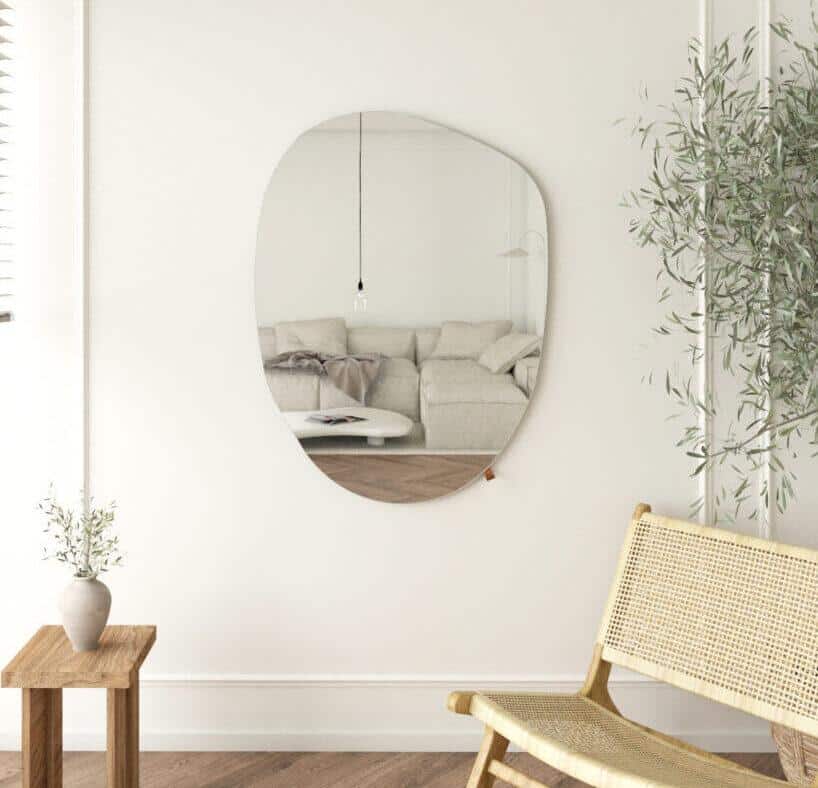 Nowoczesny salon z jasną sofą odbijającą się w lustrze ściennym o nieregularnym kształcie Lustro-japandi-110x80-cm-1-e1654706948242-1.jpg, a do tego plecione krzesło, mały drewniany stolik z wazonem, wysoka roślina narożna i minimalistyczny neutralny wystrój.